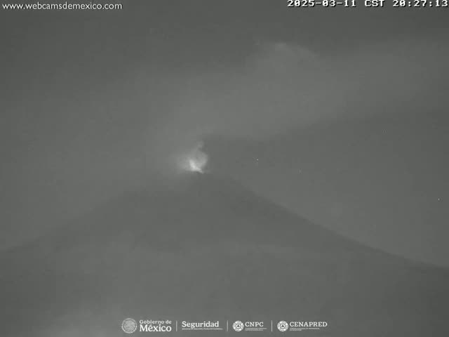 A nighttime, low-light image shows a volcano emitting a faint glow from its summit under a cloudy sky.
