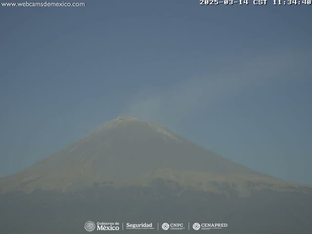 A large, snow-capped volcano is seen on a clear day with a small plume of smoke emanating from its peak.