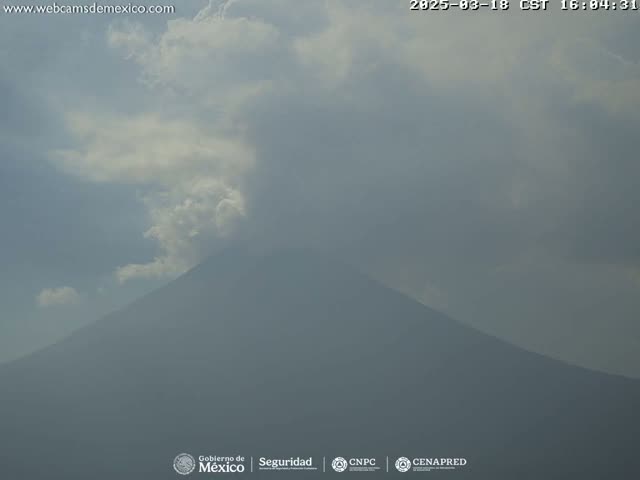 A hazy, partly cloudy sky hangs over a large, dark mountain, possibly a volcano, emitting a plume of light-colored smoke or steam.