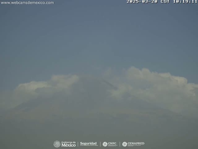 A hazy mountaintop is partially obscured by a layer of fluffy white clouds under a mostly clear, light-blue sky.
