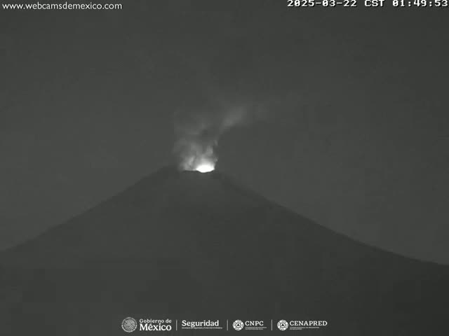 A nighttime, low-light image shows a volcano erupting with a small amount of smoke and a bright glow emanating from its crater.