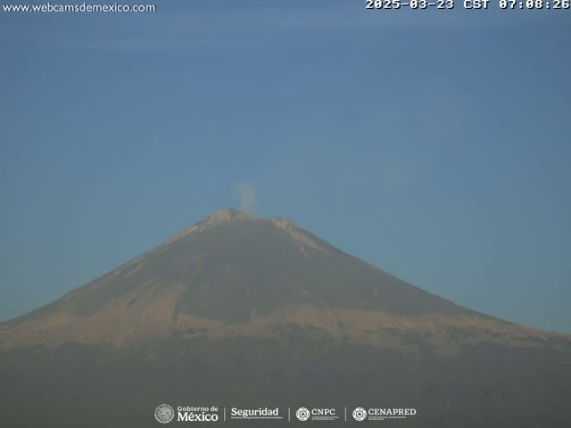 A large volcano with a small plume of smoke erupting from its peak is visible under a clear blue sky.