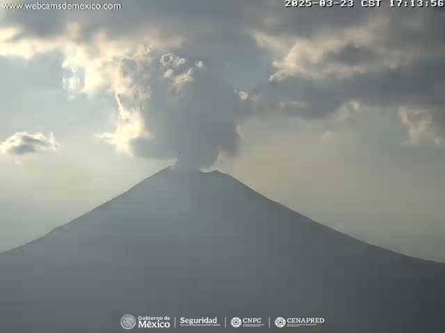 A large, dark mountain is seen emitting a plume of light gray smoke under a partly cloudy sky.