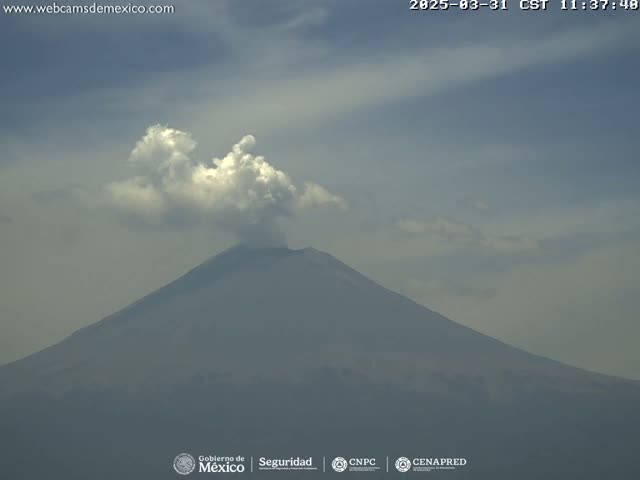 A large, dark-colored volcano with a plume of white smoke erupting from its peak is visible under a mostly cloudy sky.
