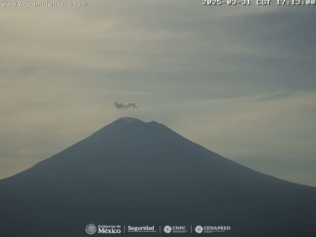 A snow-capped volcano is seen under a mostly clear, light-grey sky with a small plume of smoke emanating from its peak.