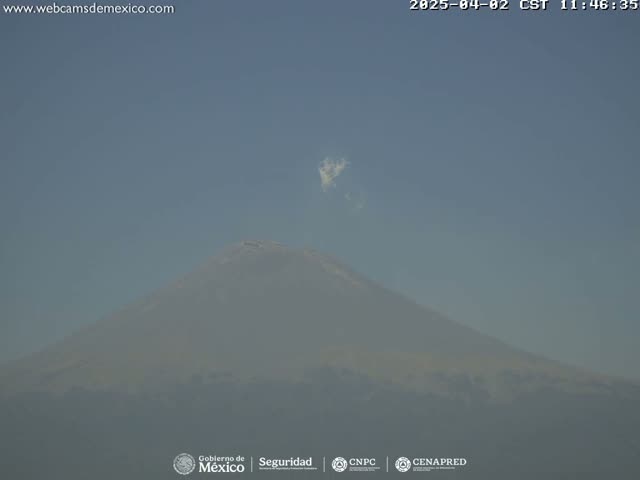 A hazy, light-blue sky shows a large, snow-capped mountain with a small plume of smoke or cloud emanating from its peak.
