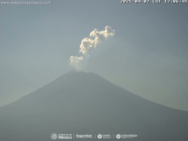 A hazy plume of smoke rises from a large, dark-grey volcano under a clear, light-grey sky.
