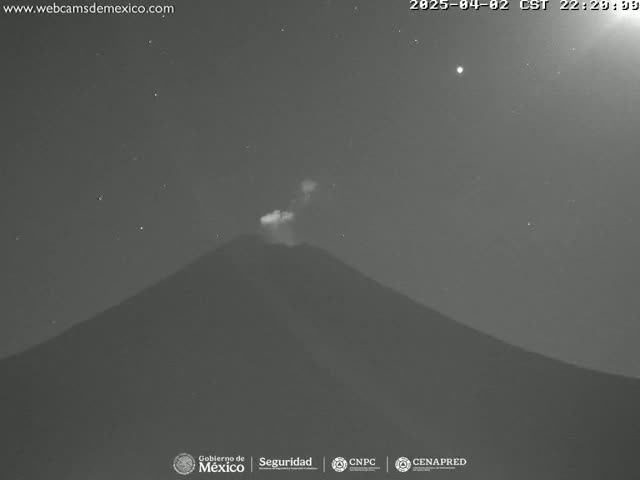 A nighttime, low-light image shows a volcano emitting a small plume of smoke or ash under a clear, starry sky.