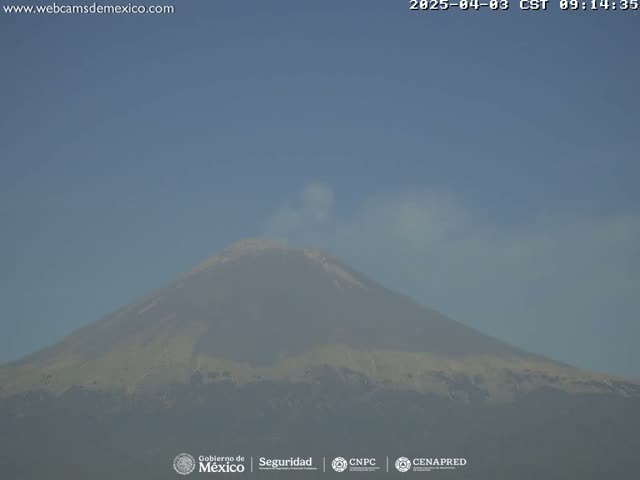 A hazy plume of smoke rises from a large, light-brown and grey volcano under a clear, light-blue sky.