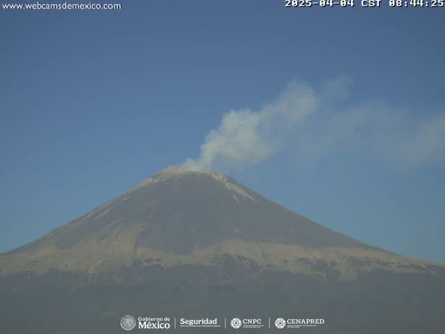 A large volcano emits a plume of light gray smoke under a clear blue sky.