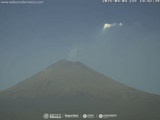 A large, tan-colored volcano is visible under a mostly clear, hazy sky with a small puff of cloud.