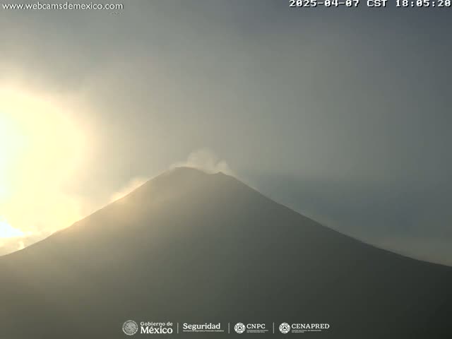 A hazy sunrise illuminates a large, dark volcano emitting a small plume of smoke.
