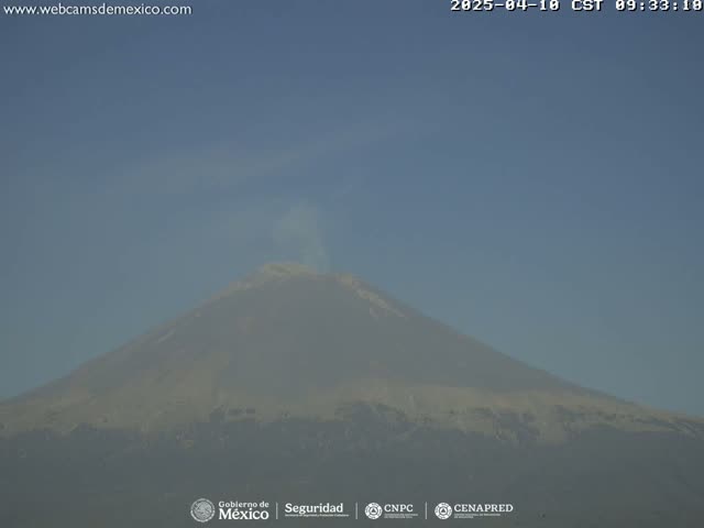 A large, hazy volcano emits a plume of smoke under a clear, light-blue sky.