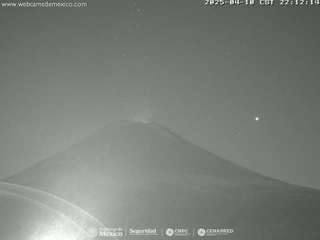 A nighttime, low-light image shows a volcano emitting a plume of smoke under a clear, starry sky.