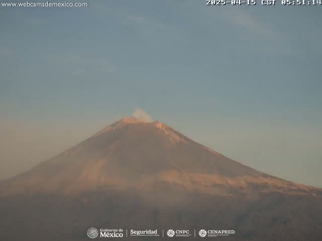 A large, dark-colored volcano with a small plume of smoke emitting from its peak is visible under a clear, light-blue sky.