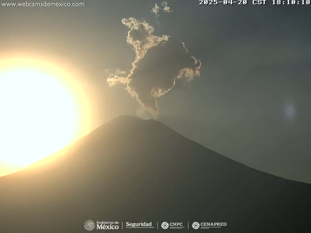 A volcano with a small plume of smoke erupting is silhouetted against a bright, setting sun in a hazy sky.