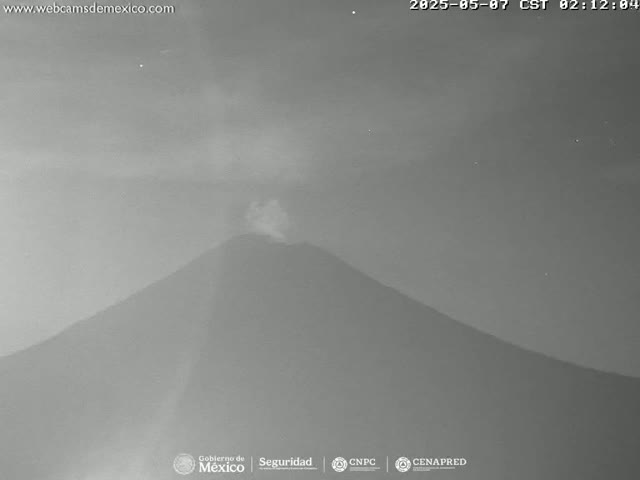 A nighttime, low-light image shows a volcano emitting a small plume of smoke under a mostly clear sky.