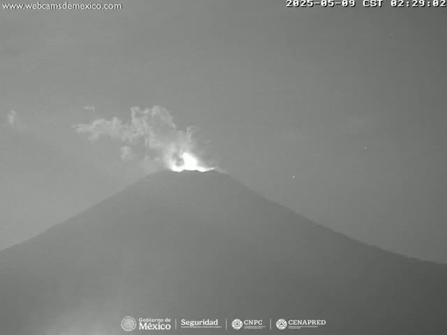 A nighttime grayscale image shows a volcano erupting with smoke and glowing lava under a mostly clear sky.