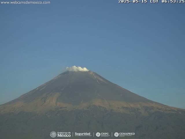 A large, dark-colored volcano with a small amount of white smoke emanating from its peak is visible under a clear, blue sky.