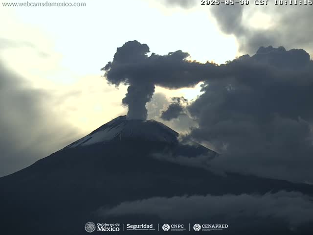 A snow-capped volcano erupts a plume of dark smoke into a partly cloudy sky.