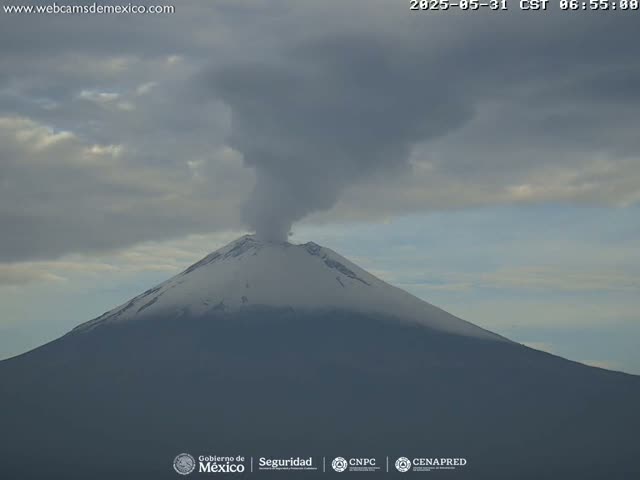 A snow-capped volcano erupts a plume of smoke under a cloudy sky.