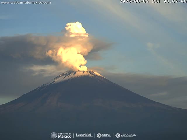 A snow-capped volcano erupts a plume of light-colored ash into a mostly clear sky.