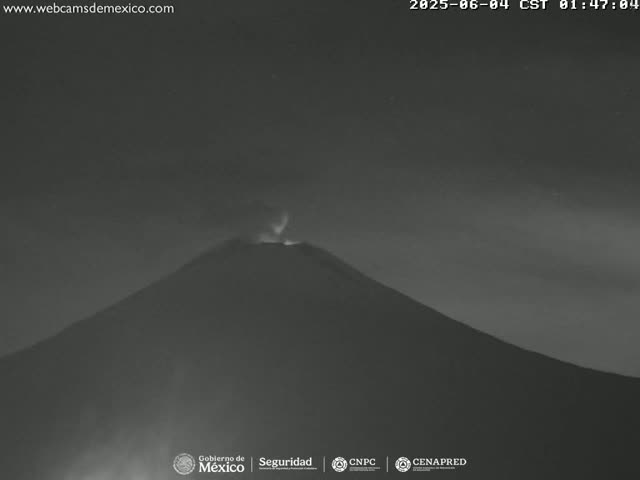 A dark, nighttime image shows a volcano emitting a faint plume of smoke or ash under a mostly clear sky.