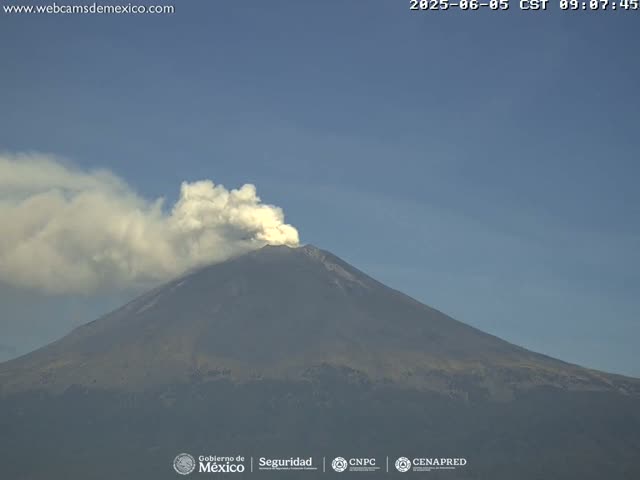 A large volcano is emitting a plume of white smoke under a clear blue sky.