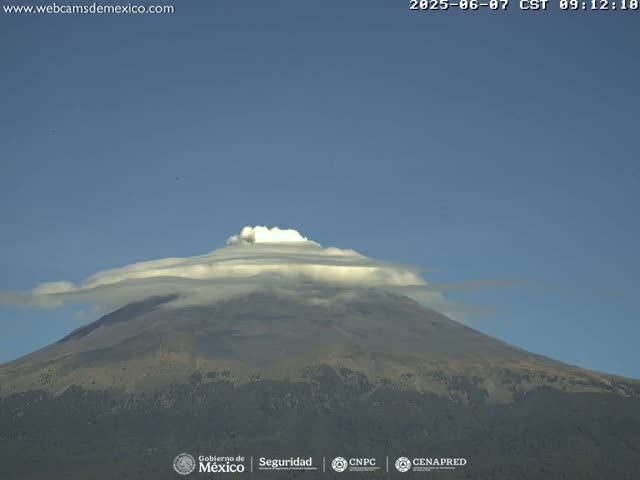 A large, dark-colored mountain is topped with a layer of white clouds under a clear blue sky.