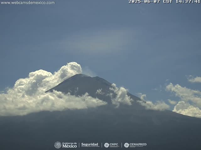 A snow-capped mountain is partially obscured by fluffy white clouds under a mostly clear blue sky.