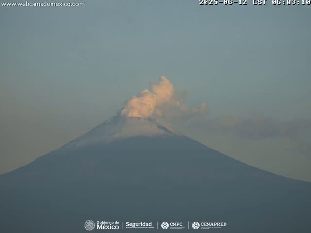 A large, dark-grey volcano is seen emitting a plume of light-colored smoke under a clear, light-grey sky.
