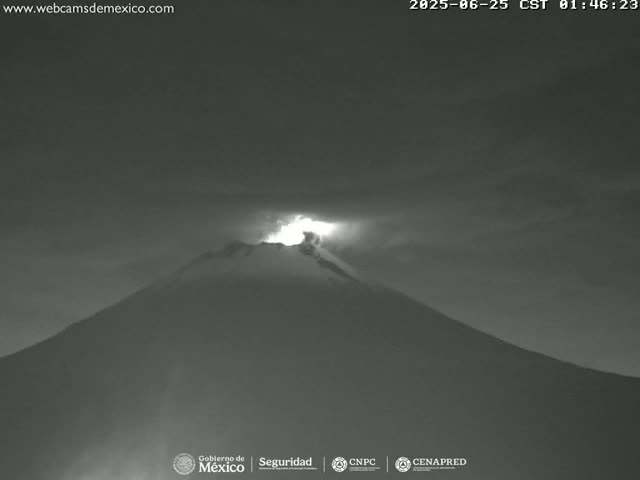 A nighttime, grayscale image shows a volcano erupting with a bright glow at its peak under a dark sky.