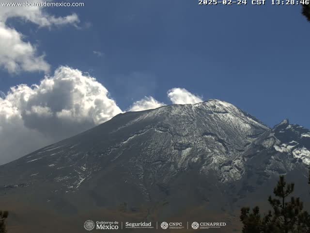A snow-capped mountain is partially obscured by fluffy white clouds under a clear blue sky.