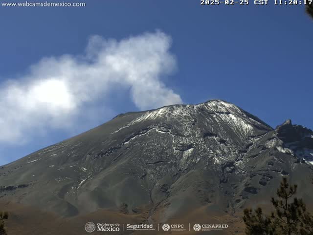 A snow-capped volcano emits a plume of white smoke under a clear blue sky.