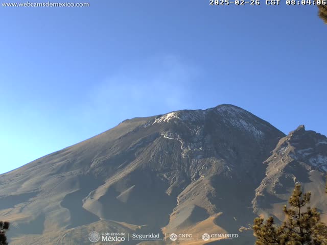 A large, dark-grey volcanic mountain with a small amount of snow near its peak is visible under a clear blue sky.