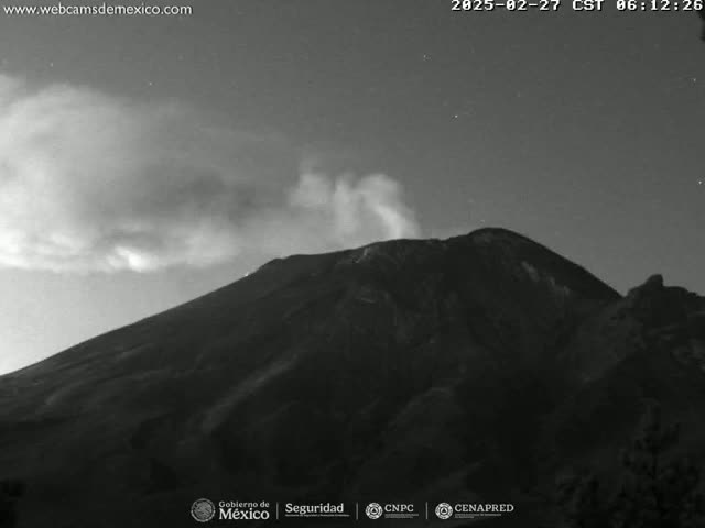 A nighttime, grayscale image shows a volcano emitting a plume of smoke under a partly cloudy sky.