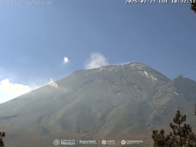 A large, dark-grey volcano with a light dusting of snow on its peak emits a small plume of light-grey smoke under a clear blue sky.