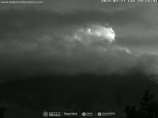 A grayscale image shows a mostly cloudy night sky over a dark, mountainous landscape.