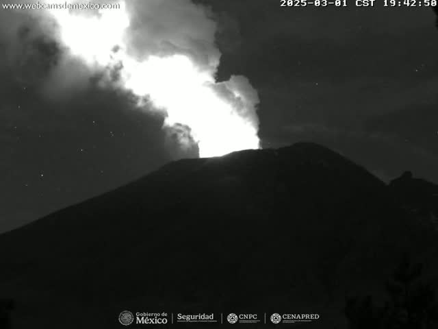 A nighttime, black and white image shows a volcano erupting with a bright plume of smoke and ash under a starry sky.