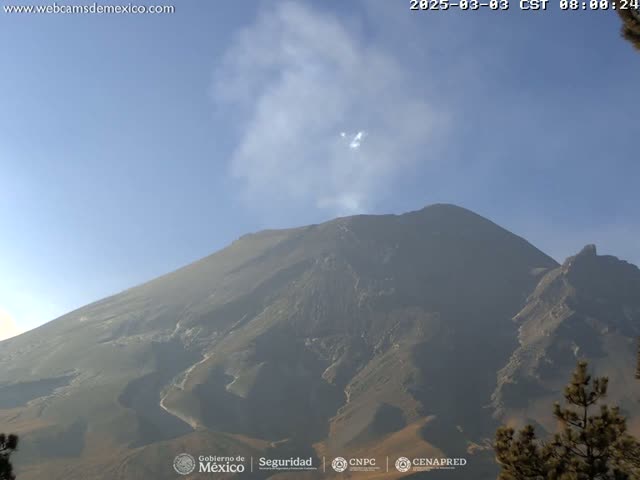 A large, dark-colored volcano emits a plume of light-grey smoke under a clear, bright blue sky.