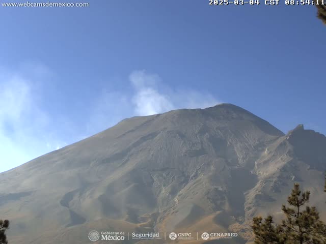 A large, dark-gray volcanic mountain is seen under a clear, bright blue sky, with a small amount of light-colored smoke emanating from its peak.