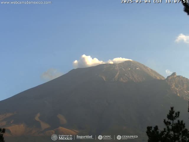 A large, dark-colored volcano is visible under a mostly clear, light-blue sky, with a small plume of white smoke emanating from its peak.