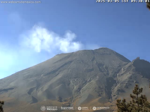 A large, dark gray volcano emits a plume of white smoke under a clear blue sky.