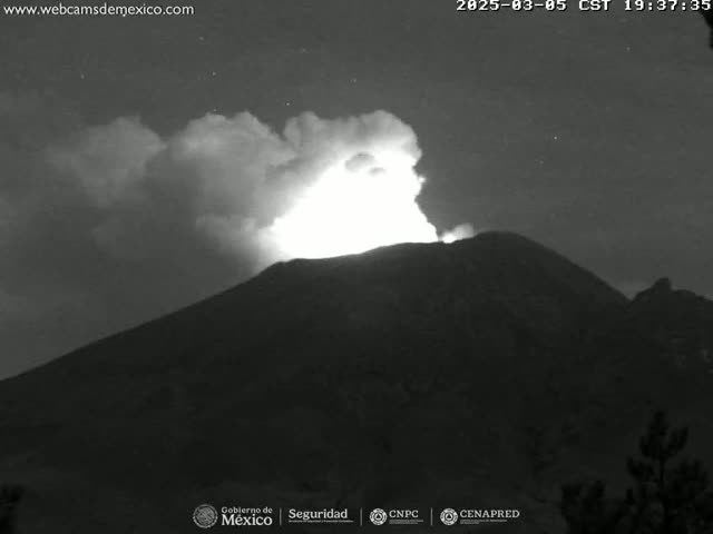 A nighttime, grayscale image shows a volcano erupting with a bright plume of ash and smoke against a dark, cloudy sky.