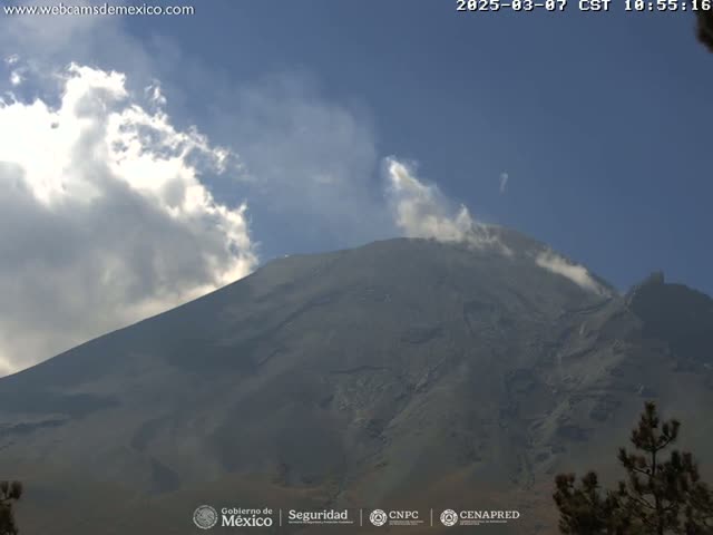 A large, dark-grey volcano with a small amount of smoke emitting from its peak is visible under a partly cloudy sky.