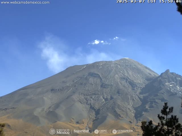 A large, gray volcano with a plume of white smoke emanating from its peak is visible under a clear blue sky.