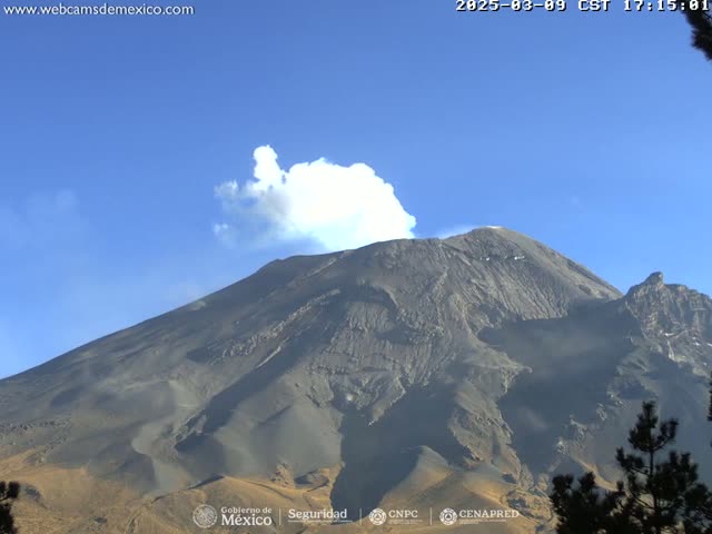 A large, dark-grey volcano with a plume of white smoke erupting from its peak is visible under a clear blue sky.