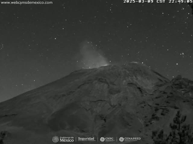 A nighttime, low-light image shows a volcano emitting a plume of smoke or ash under a starry sky.
