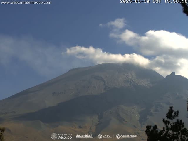 A large, dark-grey volcano is partially obscured by a thin layer of clouds under a mostly clear blue sky.