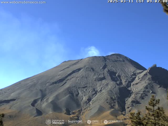 A dark gray volcanic mountain with a plume of light gray smoke billows from its peak under a clear blue sky.